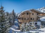 kings-avenue-luxury-chalet-courchevel-008-exterior-view-with-snow-mountains-blue-sky-and-pine-trees
